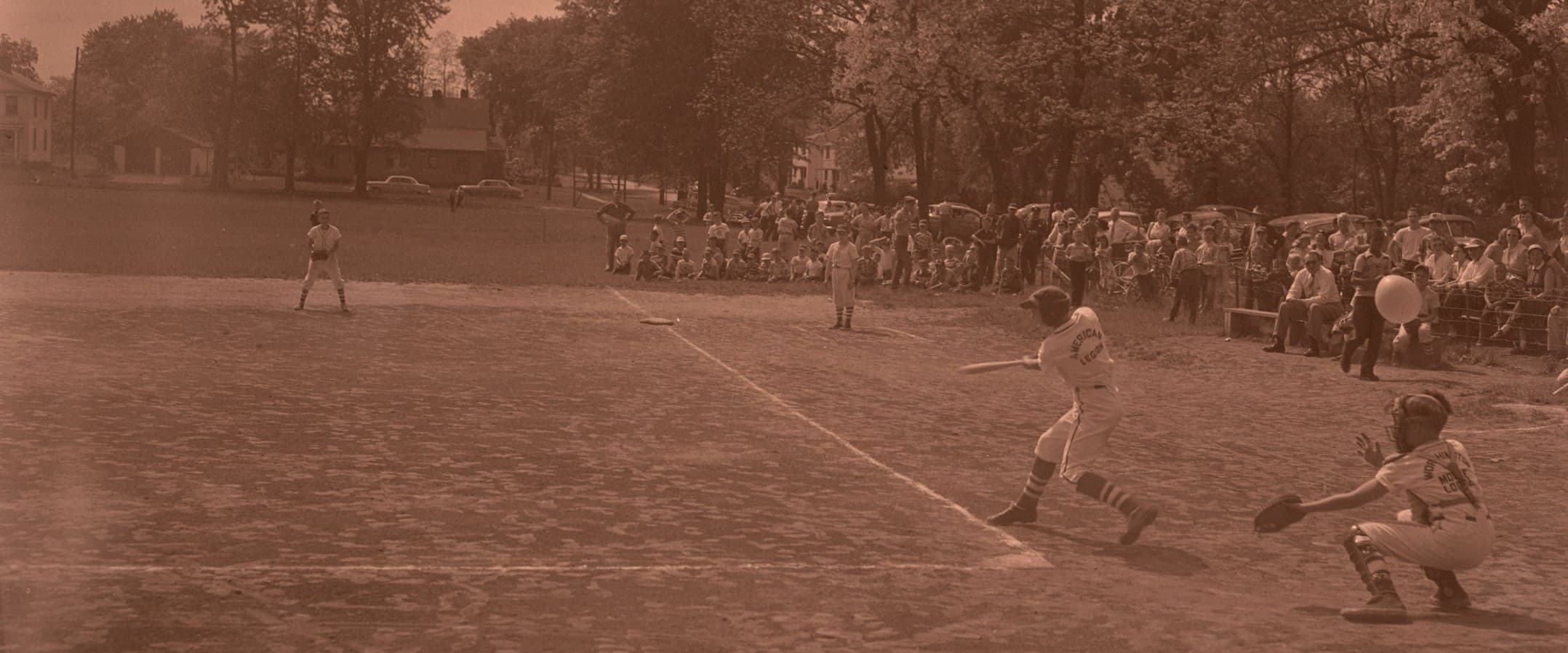 A Little League game in Hartford Park, Columbus, Ohio, around 1955: the batter mid-swing, the catcher behind him, and families watching from under the trees.